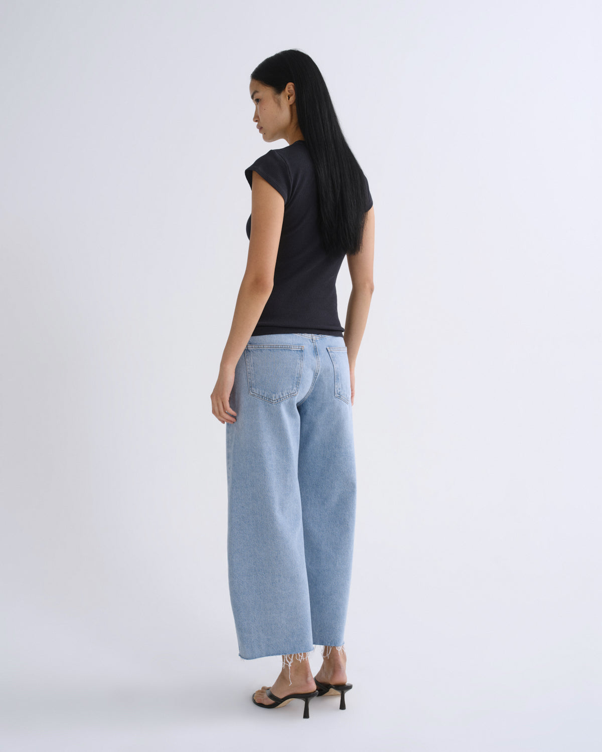 A woman shown from the back wearing a black short-sleeve top, light-wash wide-leg cropped jeans, and black heeled sandals against a plain light gray studio background.