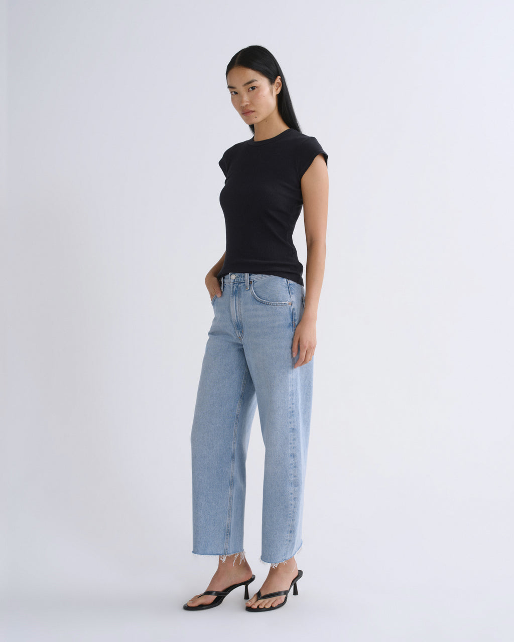 A woman modeling a black short-sleeve crewneck top and light-wash wide-leg jeans with black heeled sandals, standing against a plain light gray studio background.