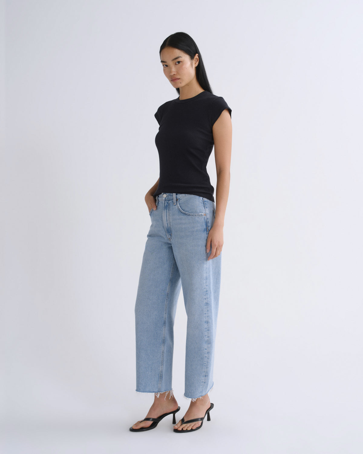 A woman modeling a black short-sleeve crewneck top and light-wash wide-leg jeans with black heeled sandals, standing against a plain light gray studio background.