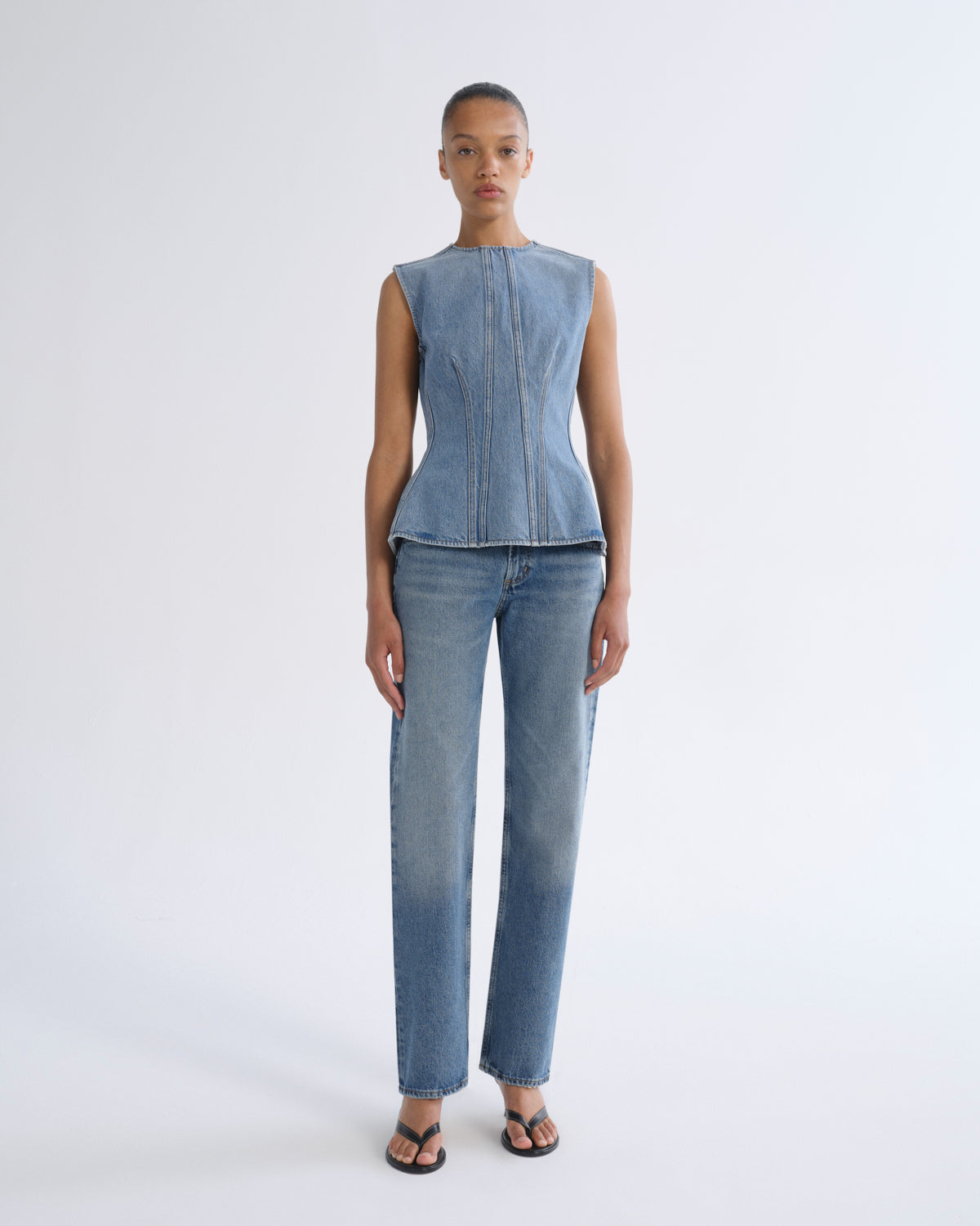 A model stands against a light gray studio background wearing a sleeveless light blue denim corset-style top and matching light blue denim straight-leg jeans with dark sandals.