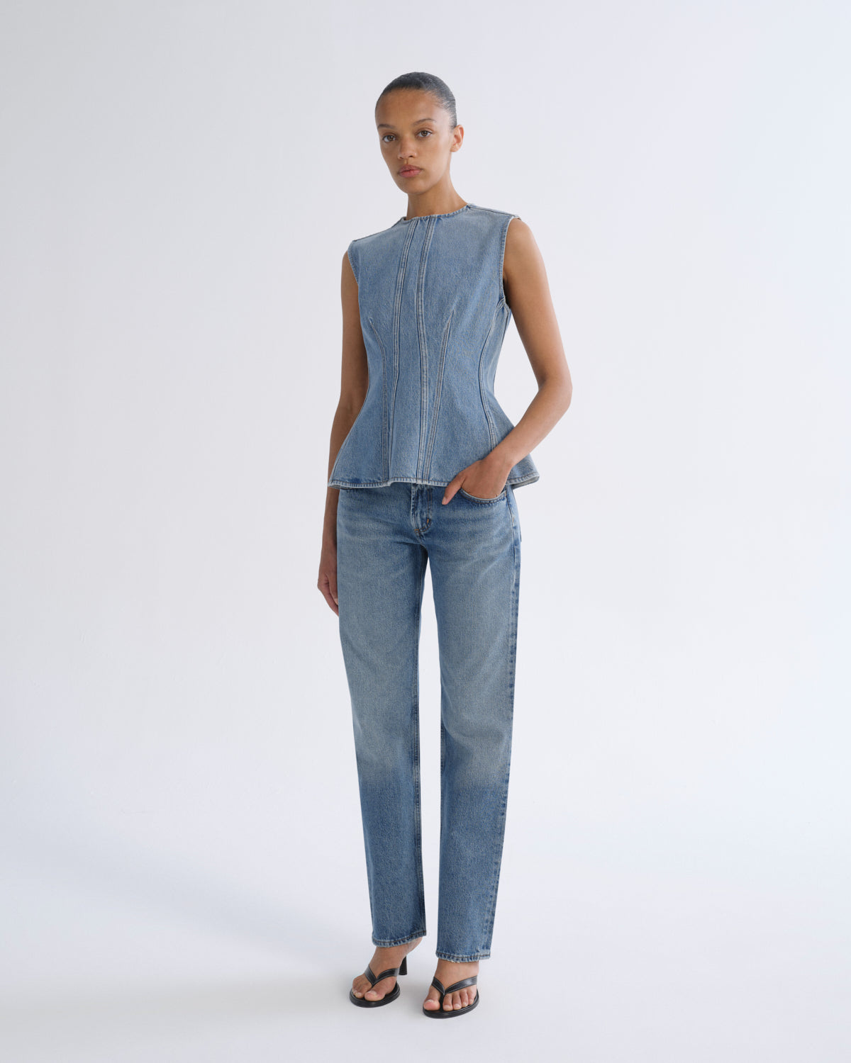 A model stands against a light gray studio background wearing a sleeveless light-wash denim top with a structured corset-style waist and matching straight-leg denim jeans.