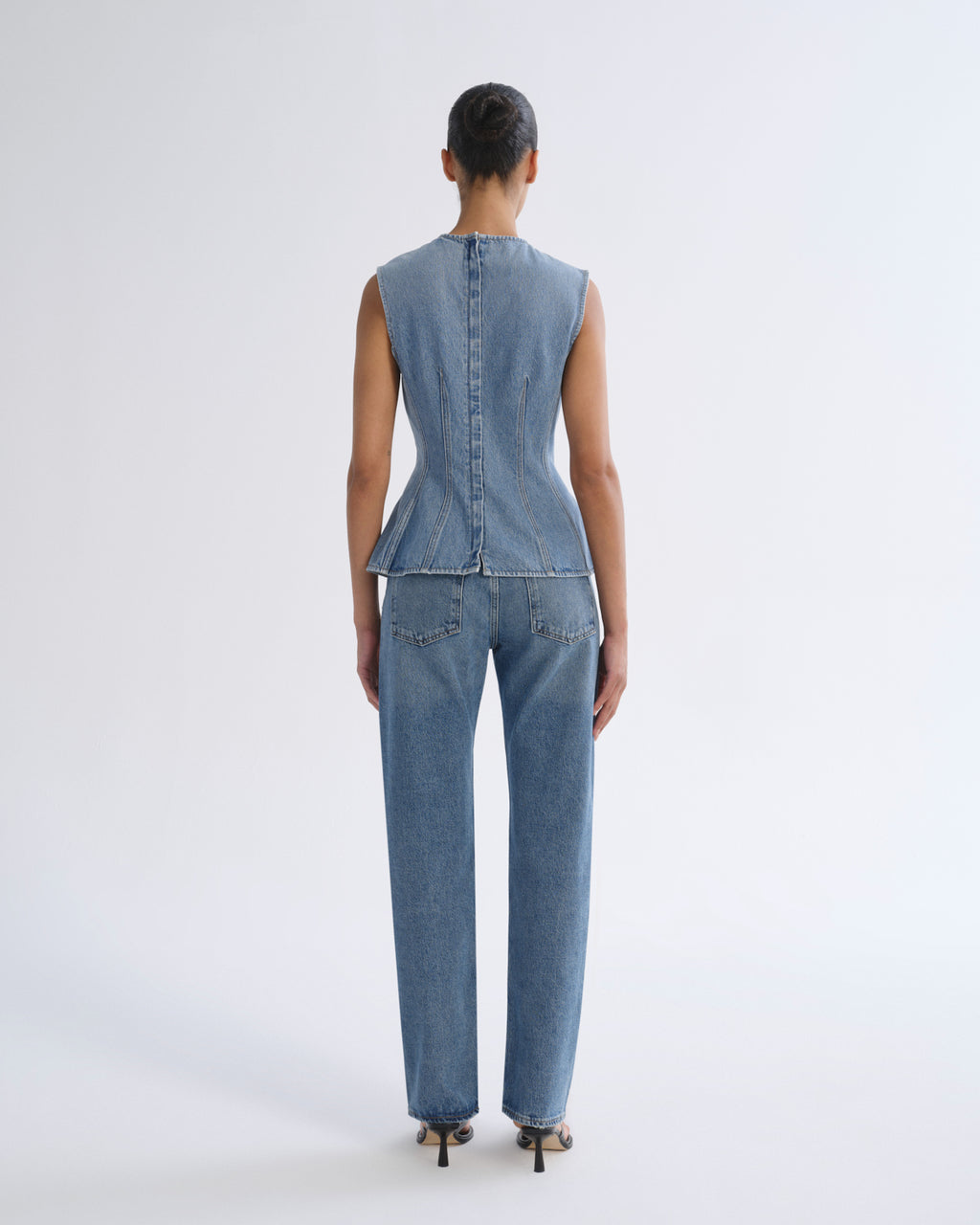 A model viewed from the back wearing a light blue denim set consisting of a sleeveless peplum-style top with a button back and matching straight-leg jeans, paired with black high heels against a light gray studio background.