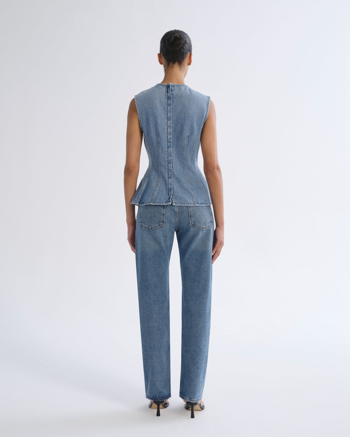 A model viewed from the back wearing a light blue denim set consisting of a sleeveless peplum-style top with a button back and matching straight-leg jeans, paired with black high heels against a light gray studio background.