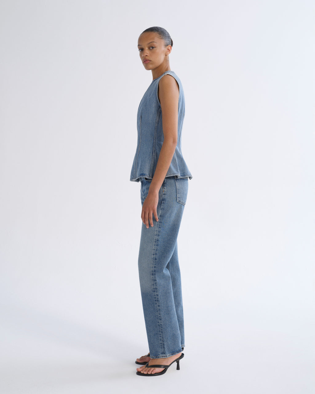 A model stands in profile wearing a sleeveless light blue denim vest and matching wide-leg jeans, paired with black heeled sandals, against a plain light gray studio background.