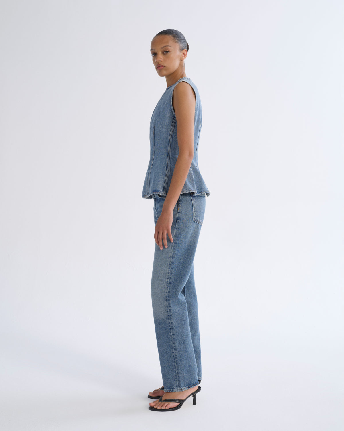 A model stands in profile wearing a sleeveless light blue denim vest and matching wide-leg jeans, paired with black heeled sandals, against a plain light gray studio background.
