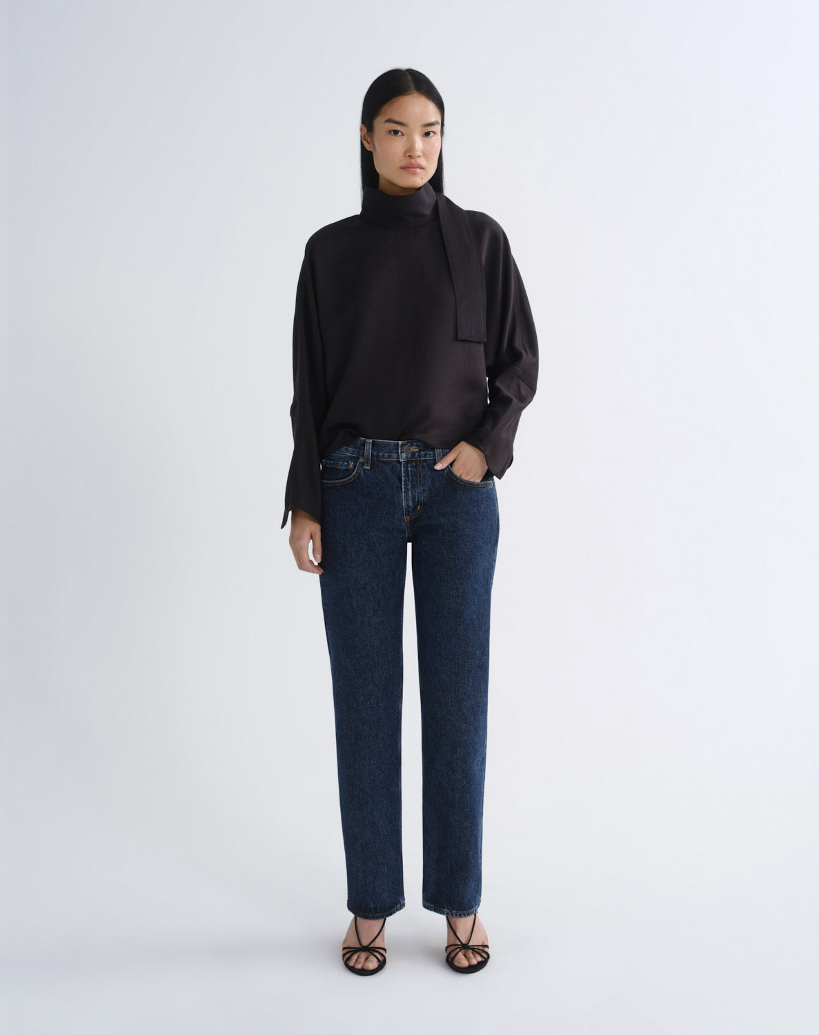 A model stands against a light gray studio background wearing a black long-sleeve mock-neck blouse with a draped collar detail, dark blue straight-leg jeans, and black strappy flat sandals.