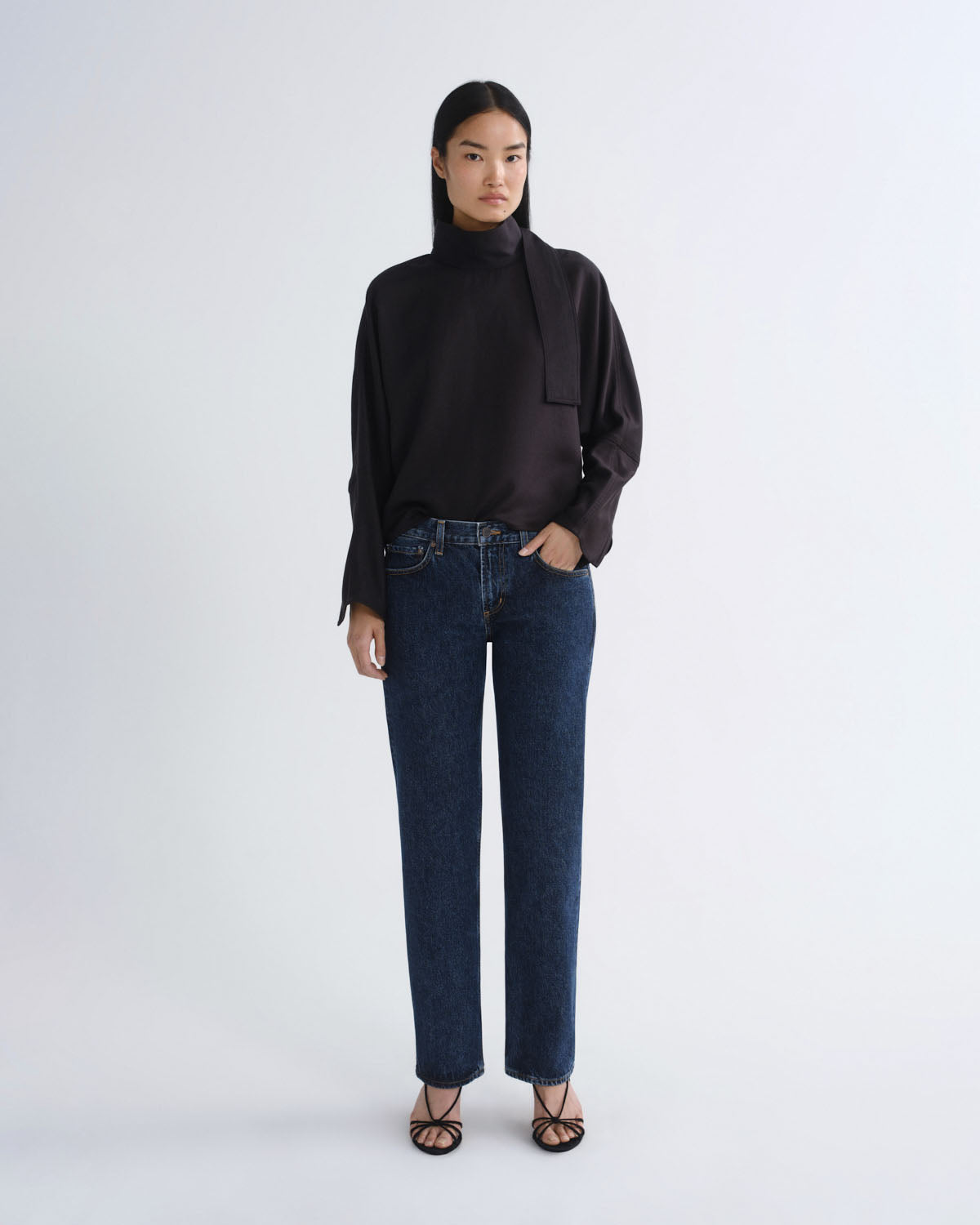 A model stands against a light gray studio background wearing a black long-sleeve mock-neck blouse with a draped collar detail, dark blue straight-leg jeans, and black strappy flat sandals.