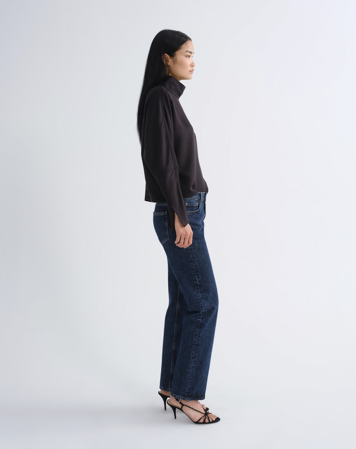 A woman stands in profile wearing a dark brown long-sleeve mock-neck top, dark blue wide-leg jeans, and black strappy high-heeled sandals against a light gray studio background.