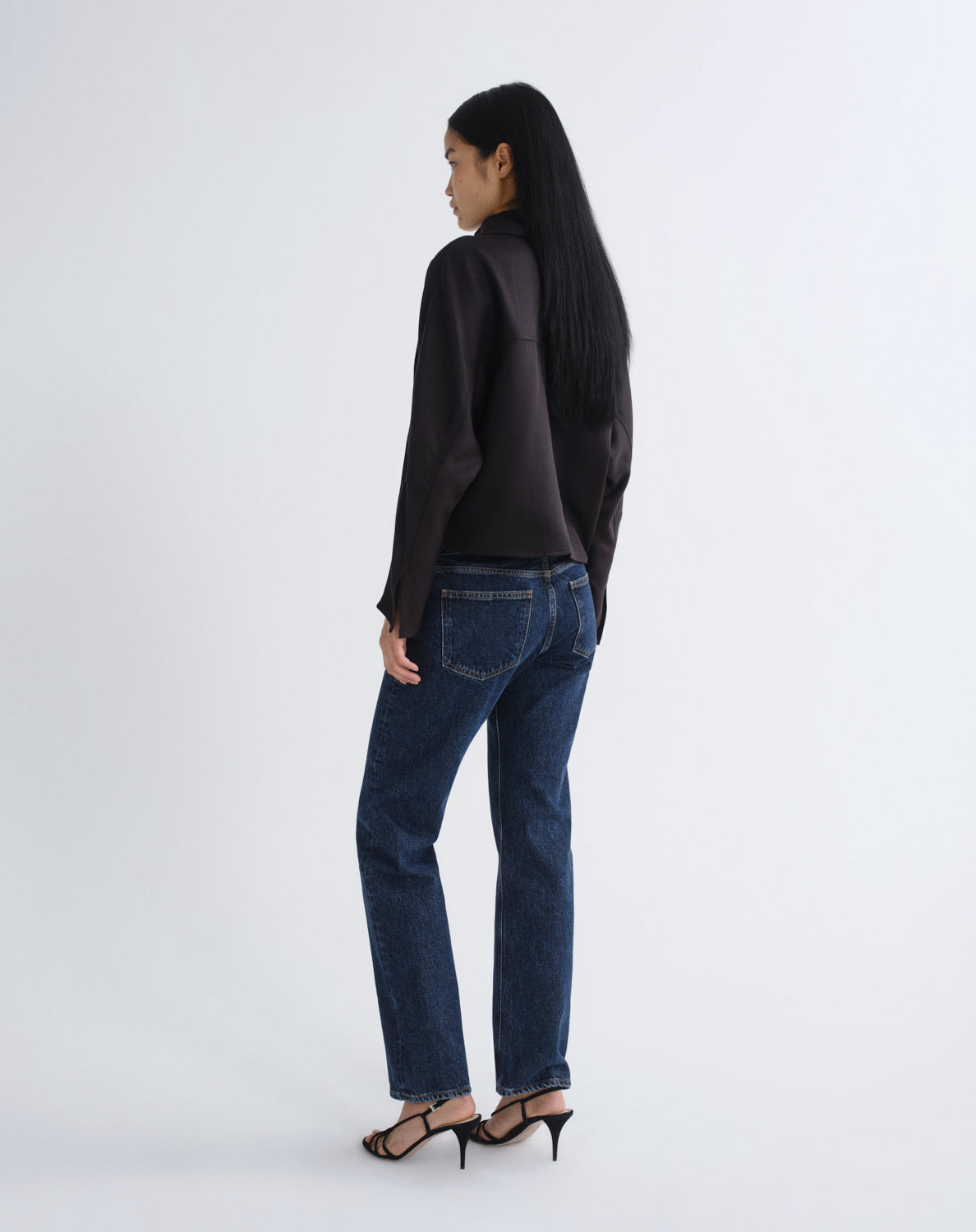 A woman is shown from the back wearing dark blue straight-leg jeans, a black long-sleeve top, and black high-heeled sandals against a plain white studio background.