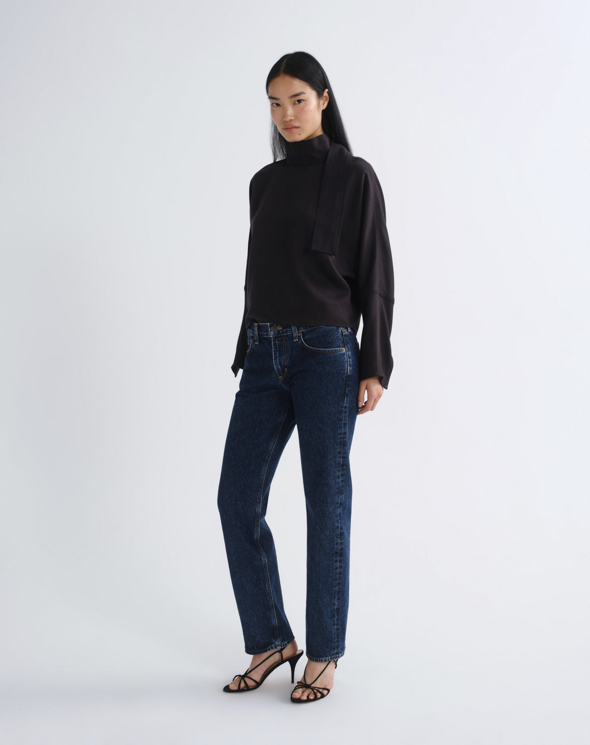 A model stands against a light gray studio background wearing a black long-sleeve mock-neck top with a draped collar detail, dark blue straight-leg jeans, and black strappy high-heeled sandals.