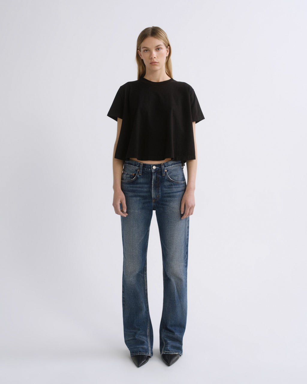 A model stands against a light gray studio background wearing a black short-sleeve cropped t-shirt and blue straight-leg jeans.