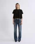 A model stands against a light gray studio background wearing a black short-sleeve cropped t-shirt and blue straight-leg jeans.