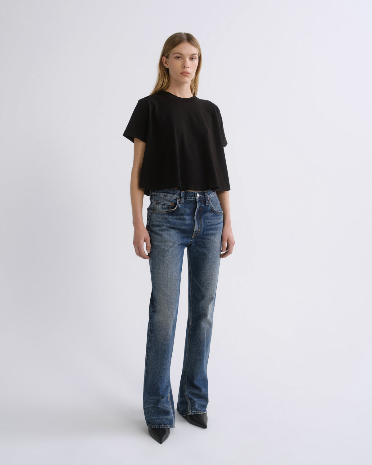 A model stands against a light gray studio background wearing a black short-sleeve cropped T-shirt and blue straight-leg jeans.