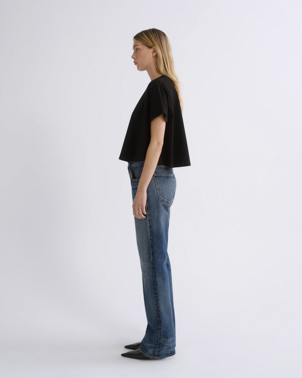 A woman stands in profile wearing a black cropped short-sleeve top and blue jeans against a plain light gray studio background.