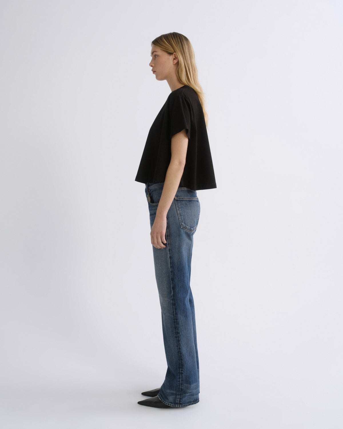 A woman stands in profile wearing a black cropped short-sleeve top and blue jeans against a plain light gray studio background.
