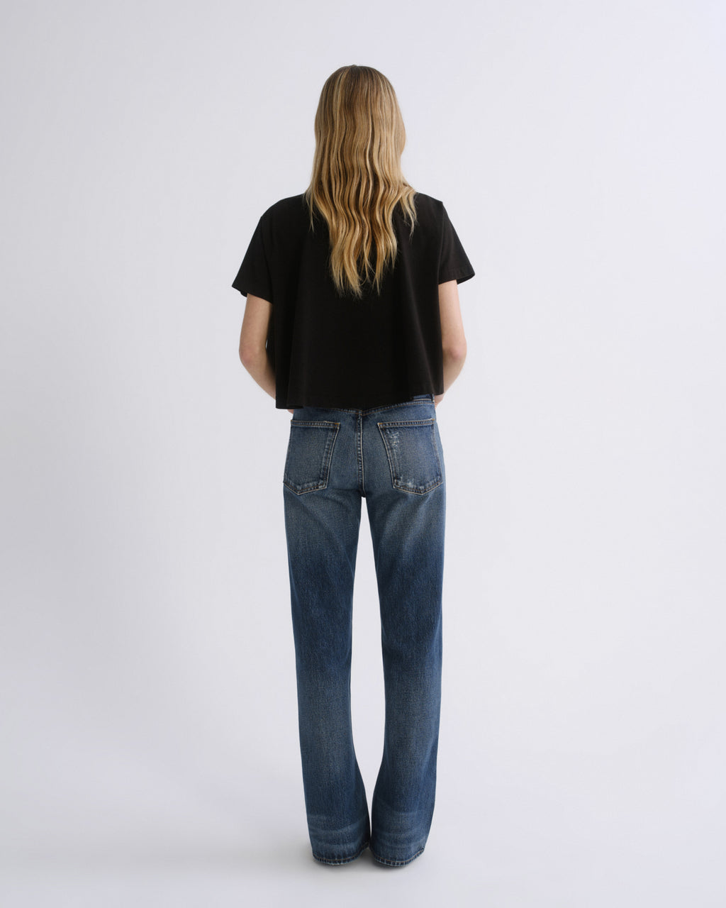 A woman viewed from behind wearing a black short-sleeve crop top and blue straight-leg jeans against a light gray studio background.
