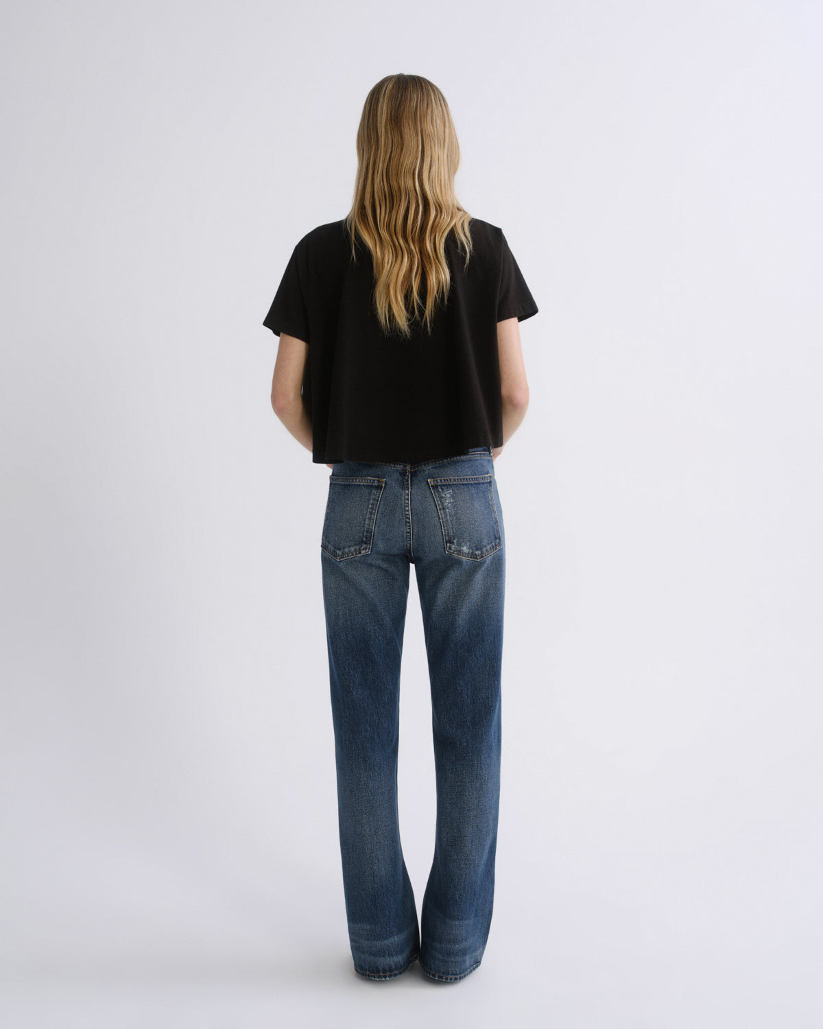 A woman viewed from behind wearing a black short-sleeve crop top and blue straight-leg jeans against a light gray studio background.