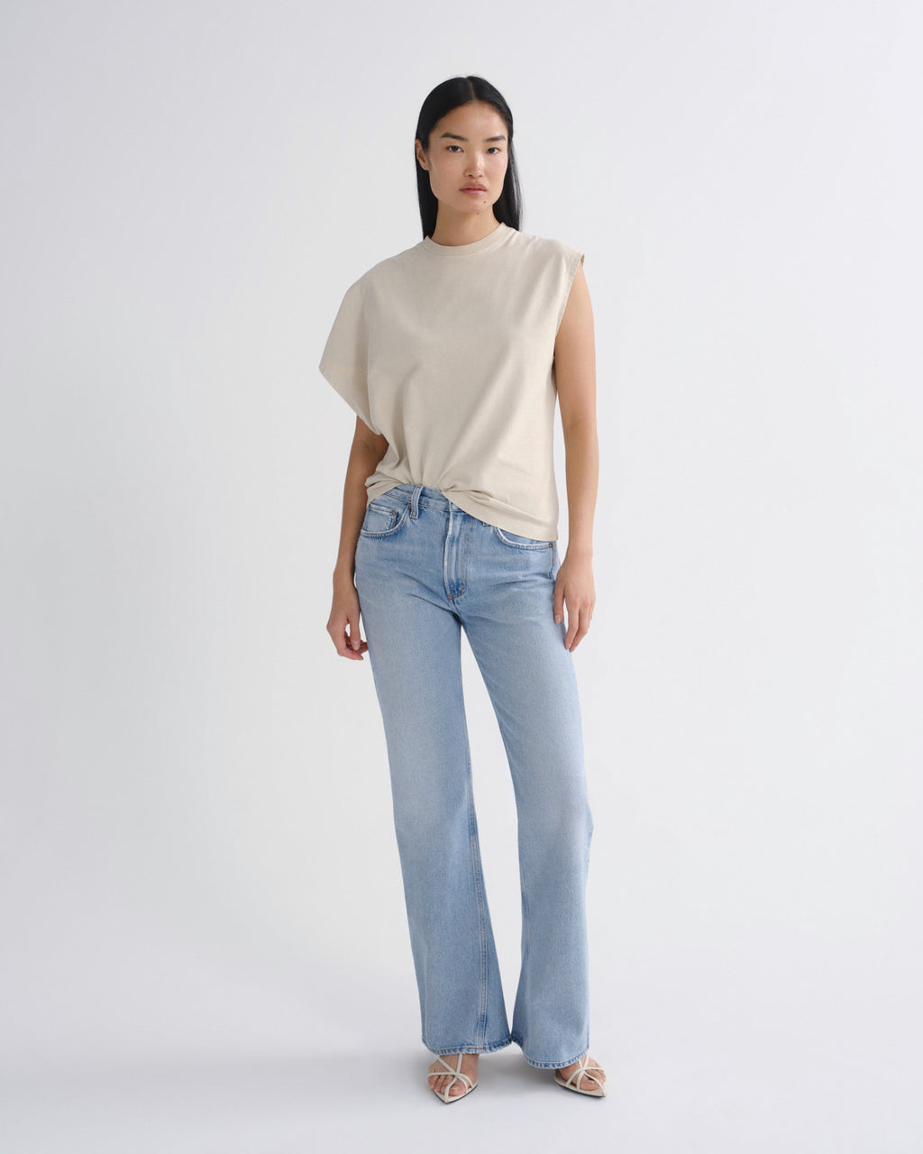 A model stands against a light gray studio background wearing a sleeveless cream top with draped side details and light-wash blue jeans.