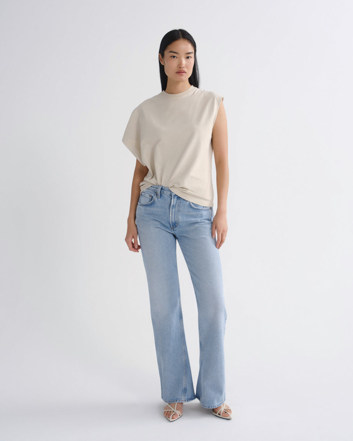A model stands against a light gray studio background wearing a sleeveless cream top with draped side details and light-wash blue jeans.