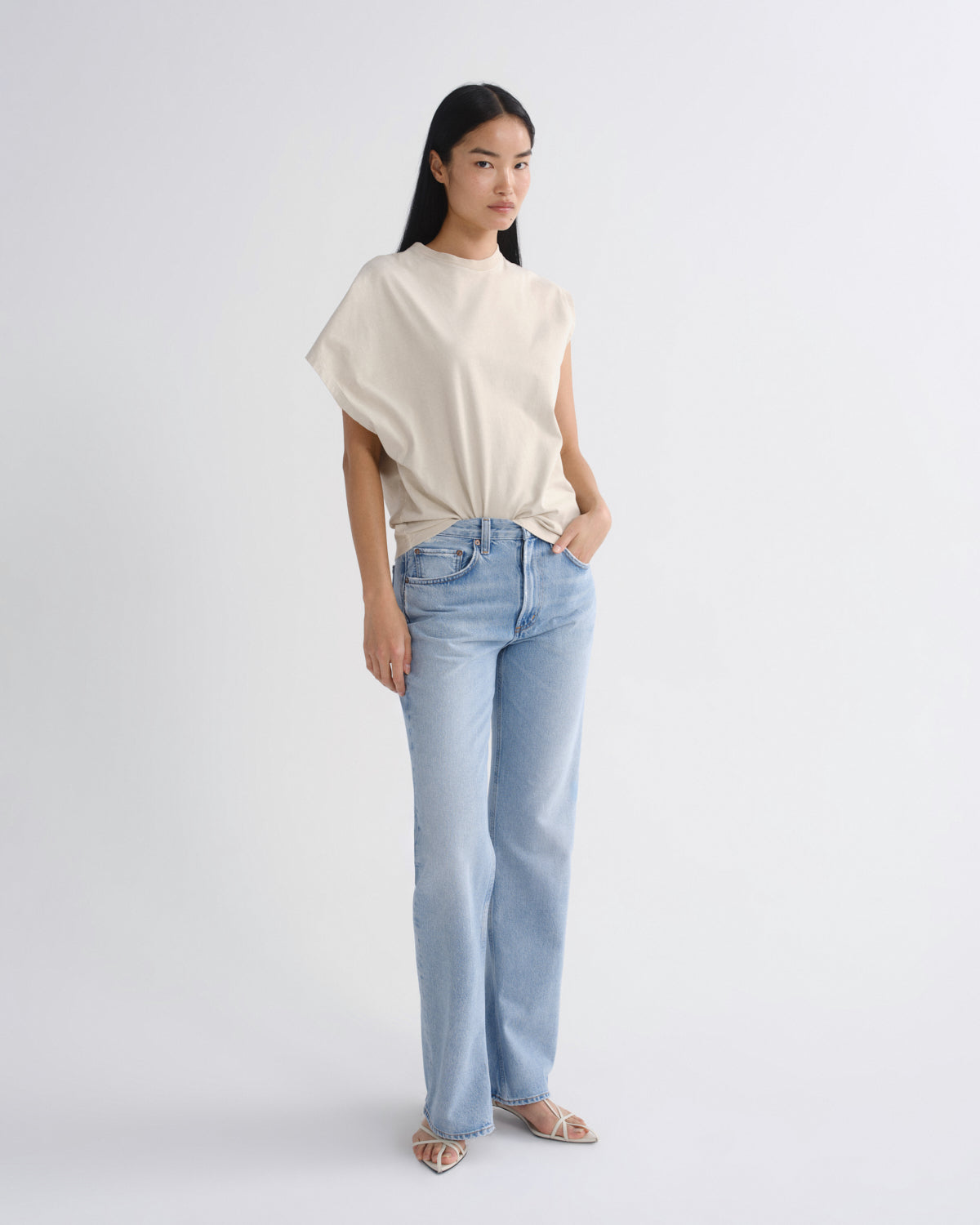 A woman modeling a cream-colored, sleeveless draped top and light-wash blue jeans against a plain white studio background.
