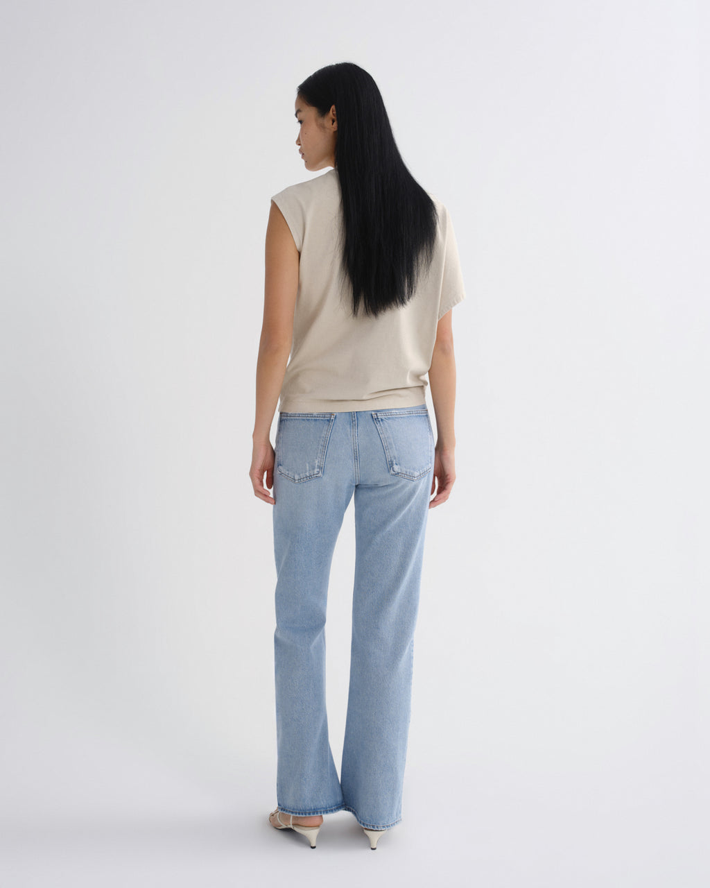 A woman with long black hair is shown from behind wearing a beige sleeveless top and light blue straight-leg jeans, standing against a plain light gray studio background.