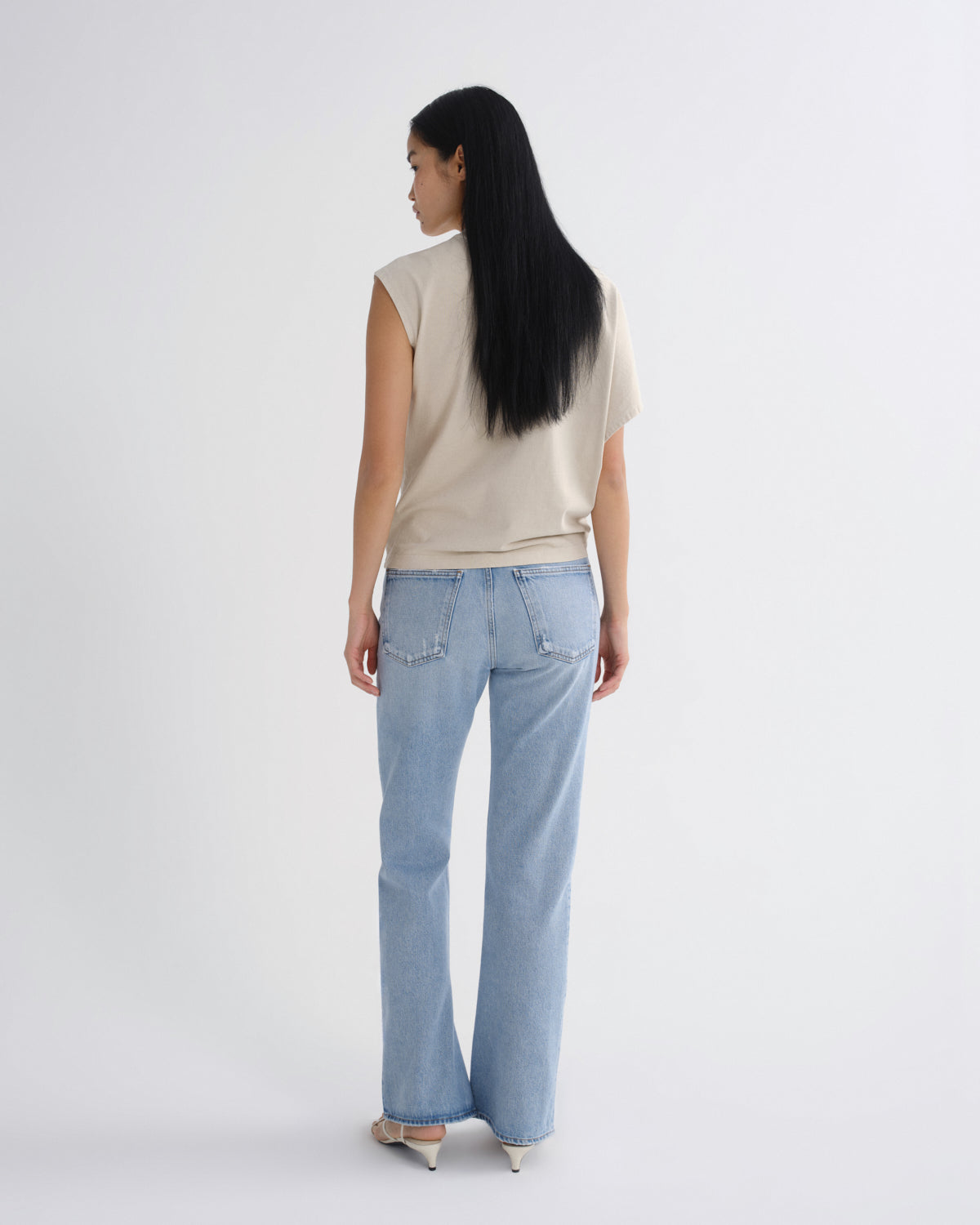 A woman with long black hair is shown from behind wearing a beige sleeveless top and light blue straight-leg jeans, standing against a plain light gray studio background.