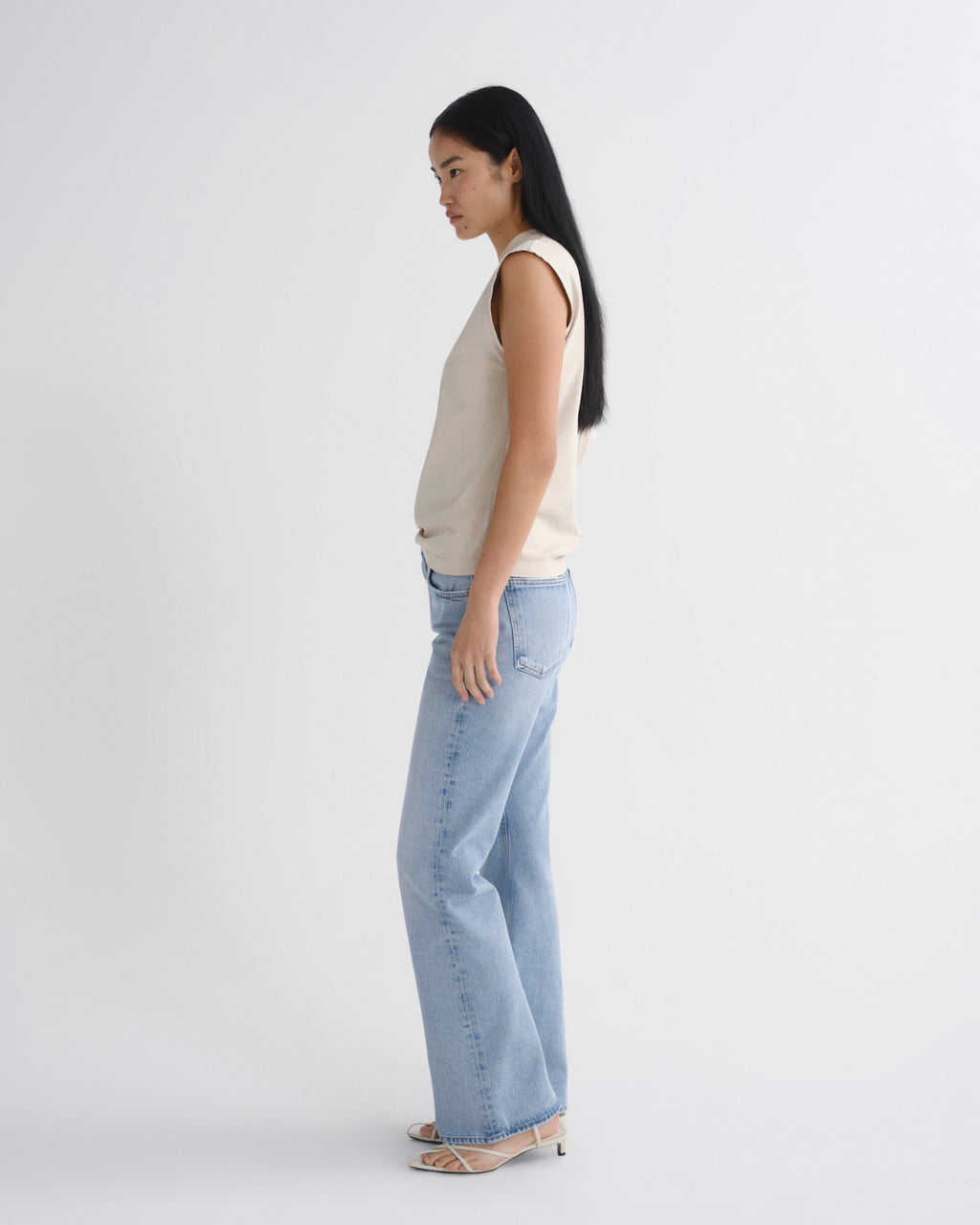 A woman stands in profile wearing a sleeveless cream top, light blue wide-leg jeans, and beige heeled sandals against a plain light gray studio background.