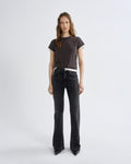 A woman stands against a plain light gray studio background wearing a dark brown short-sleeve t-shirt with white ribbed trim, black wide-leg jeans, and black pointed shoes.