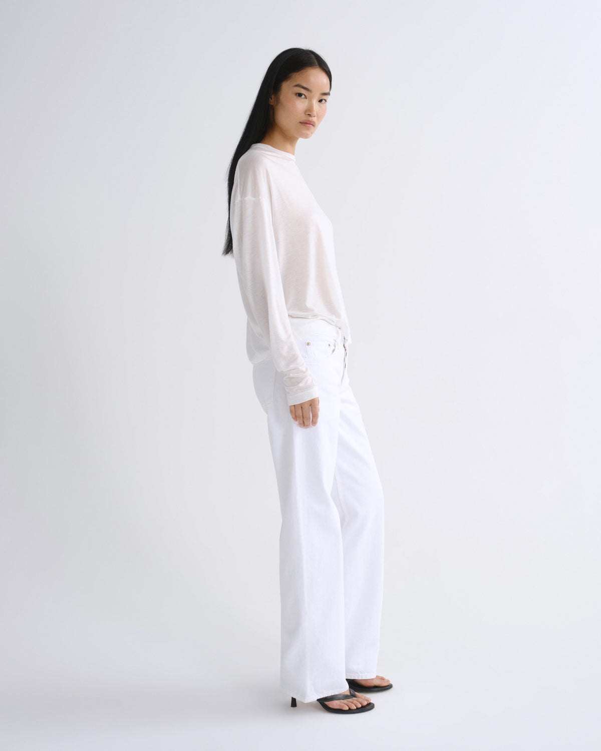 A woman modeling a white long-sleeve top and white wide-leg pants, standing in profile against a clean white studio background.