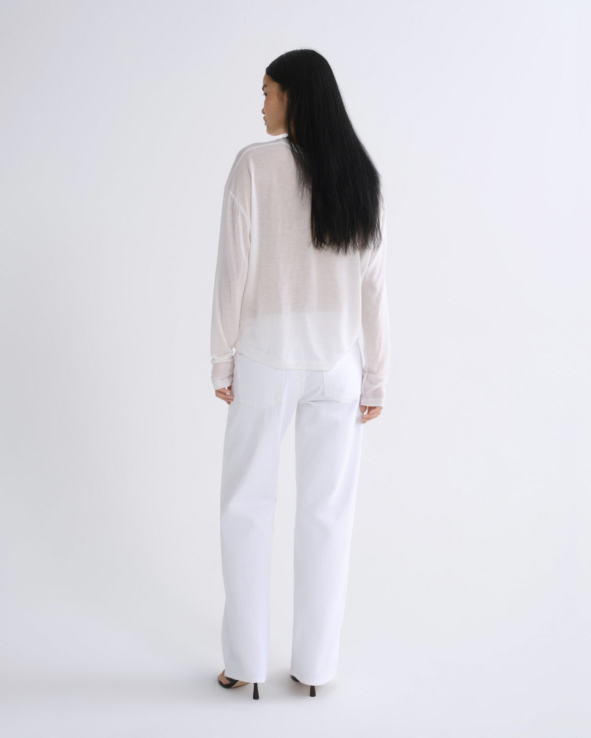 A woman with long black hair is shown from behind wearing a sheer white long-sleeve top and white wide-leg trousers against a plain white studio background.