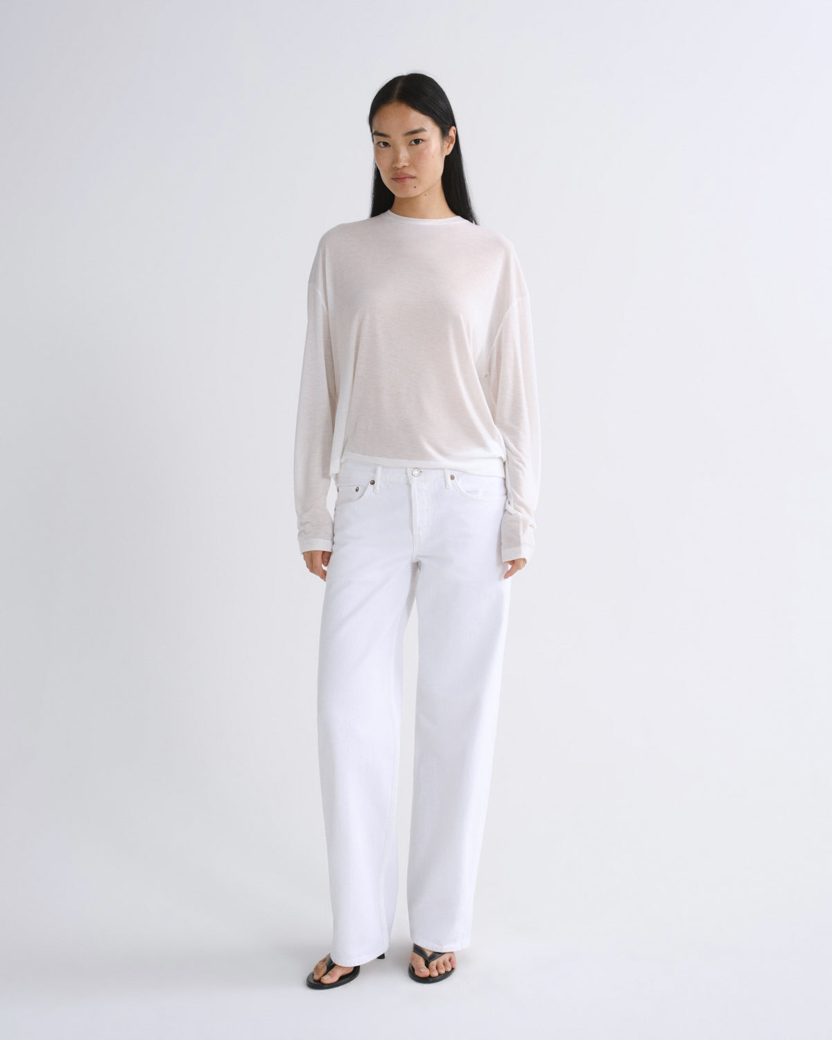 A woman modeling a sheer, long-sleeve, off-white top paired with white wide-leg jeans and black sandals, standing against a seamless light gray studio background.