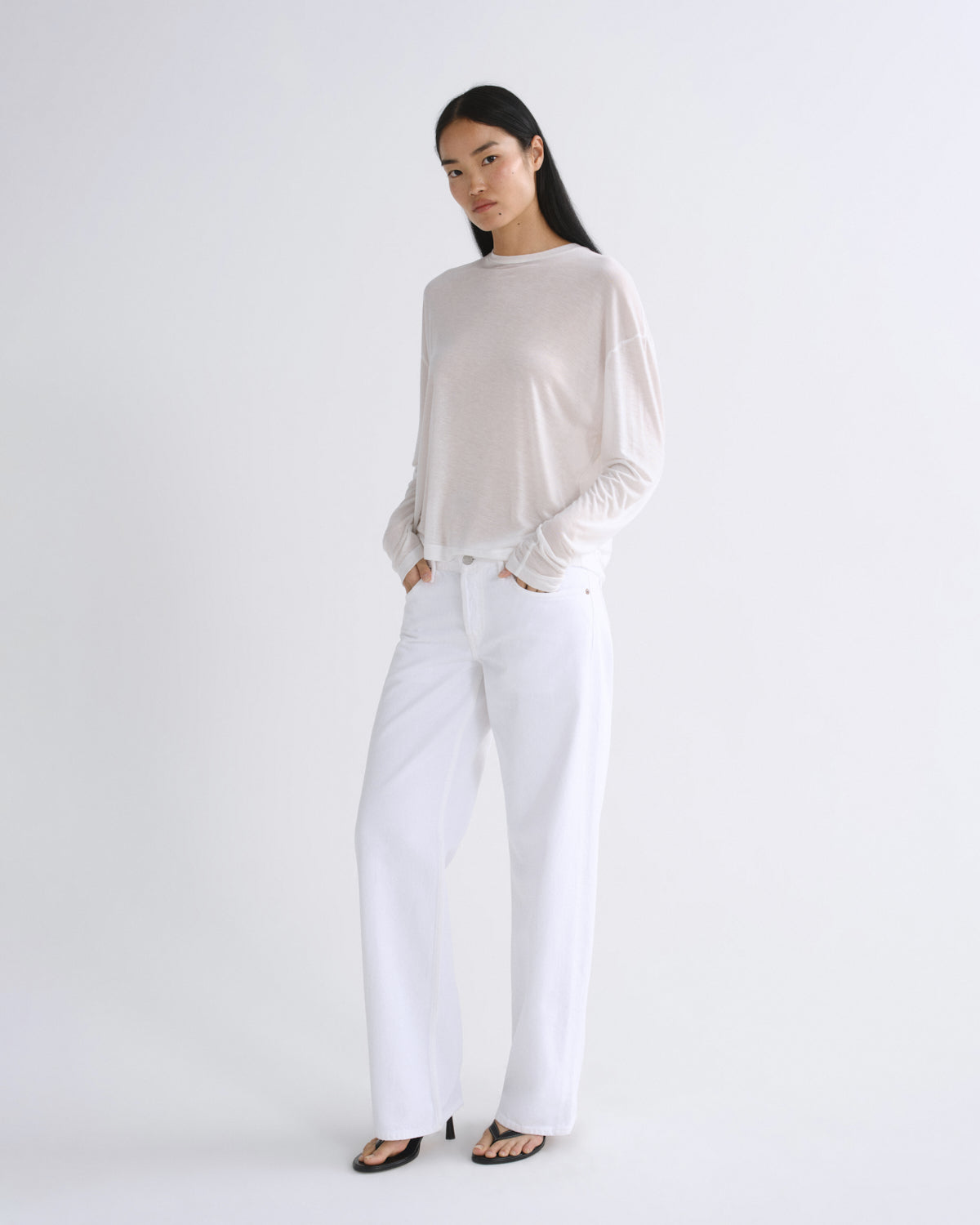 A model stands against a plain white studio background wearing a light beige long-sleeve knit top, white wide-leg pants, and black sandals.