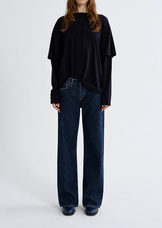 Person wearing black layered top with blue pants on a grey background
