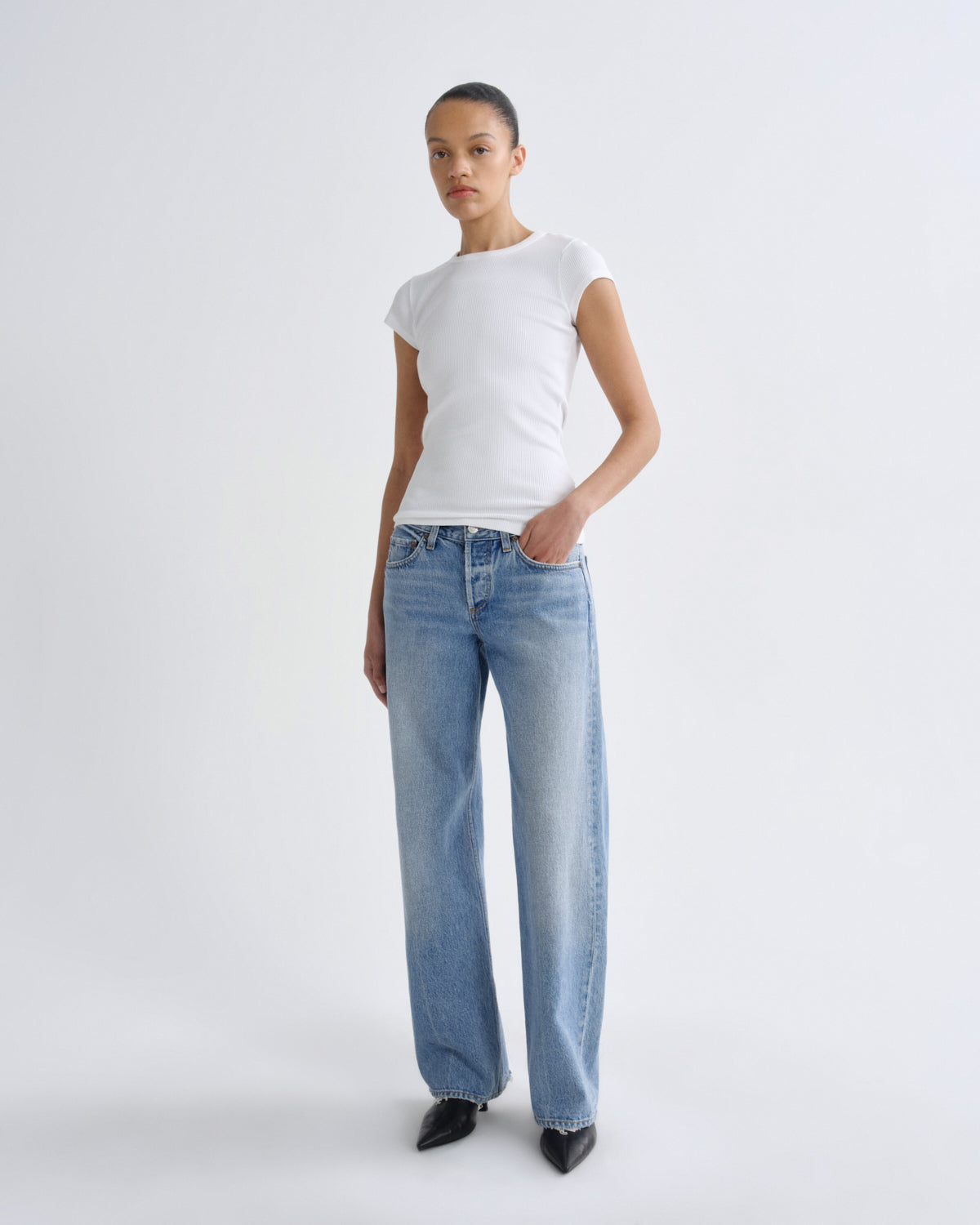 A model stands against a light gray studio background wearing a white short-sleeve crewneck t-shirt, light-wash blue wide-leg jeans, and black pointed-toe heels.
