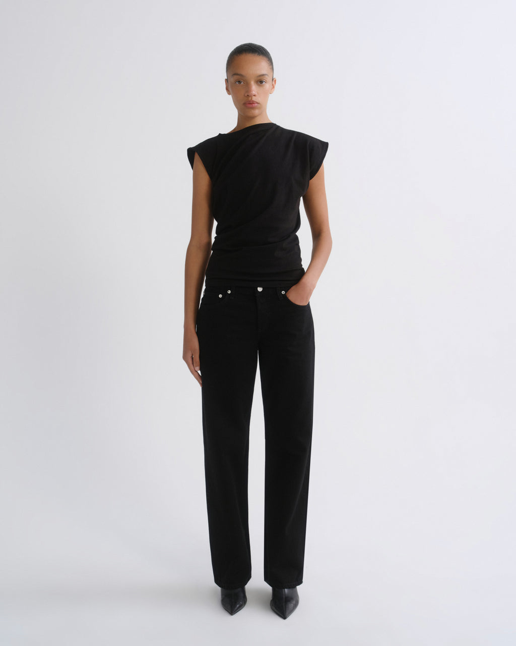 A model stands against a light gray studio background wearing a black asymmetrical draped top with cap sleeves and black wide-leg trousers.