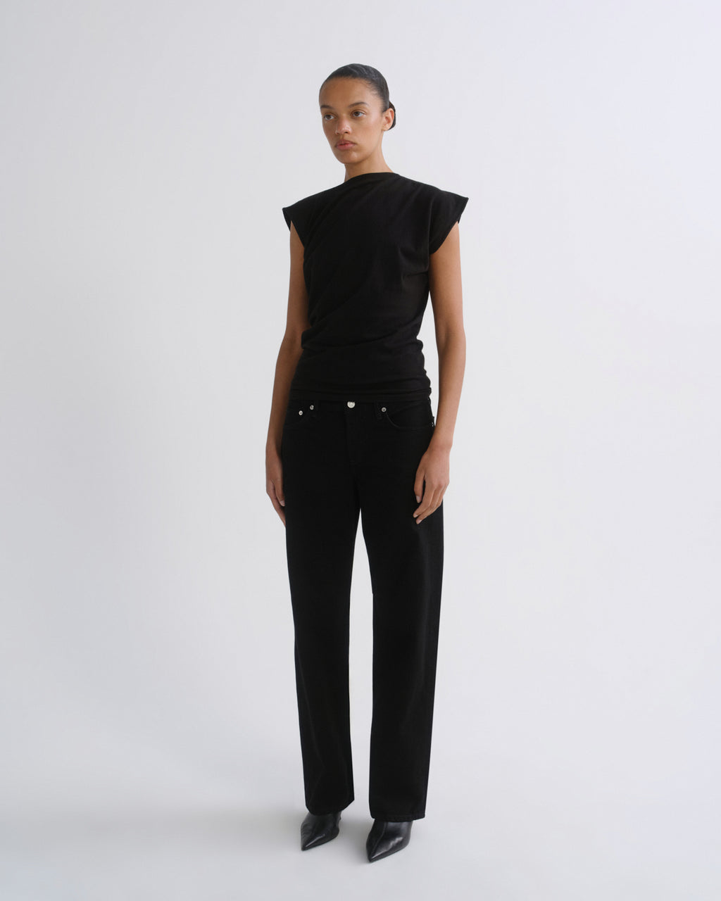 A model stands against a light gray studio background wearing a black asymmetrical short-sleeve top and black wide-leg trousers.