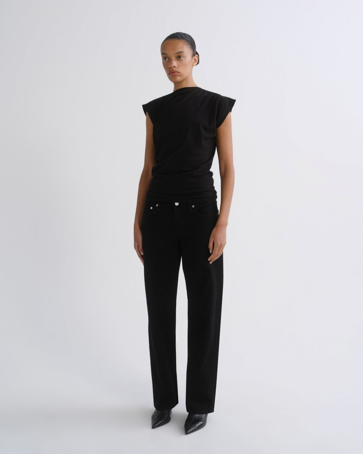 A model stands against a light gray studio background wearing a black asymmetrical short-sleeve top and black wide-leg trousers.