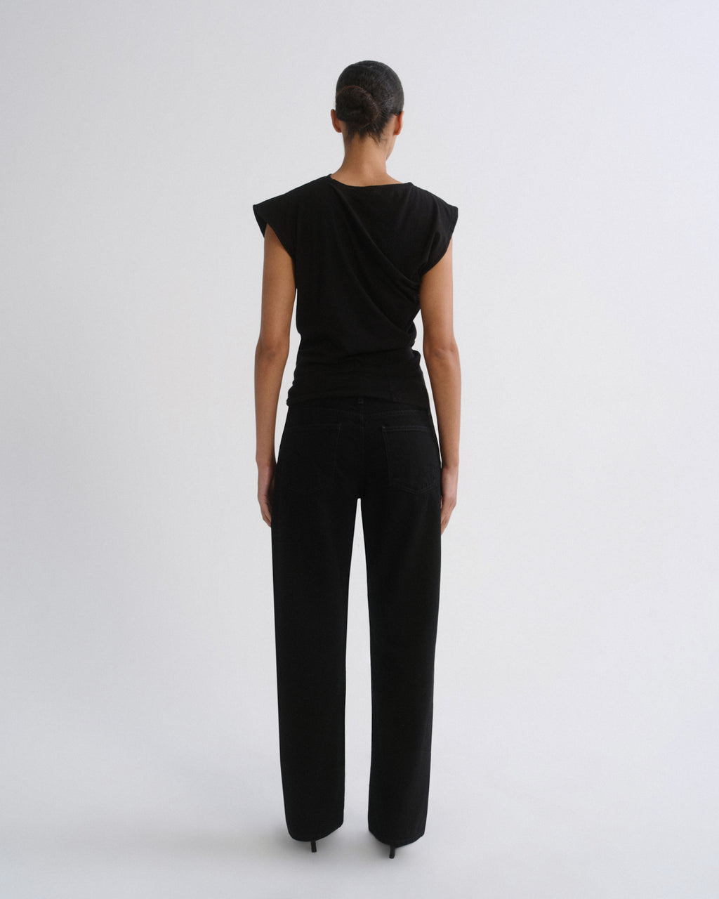 A model viewed from the back wearing a black draped sleeveless top and black wide-leg trousers against a light gray studio background.