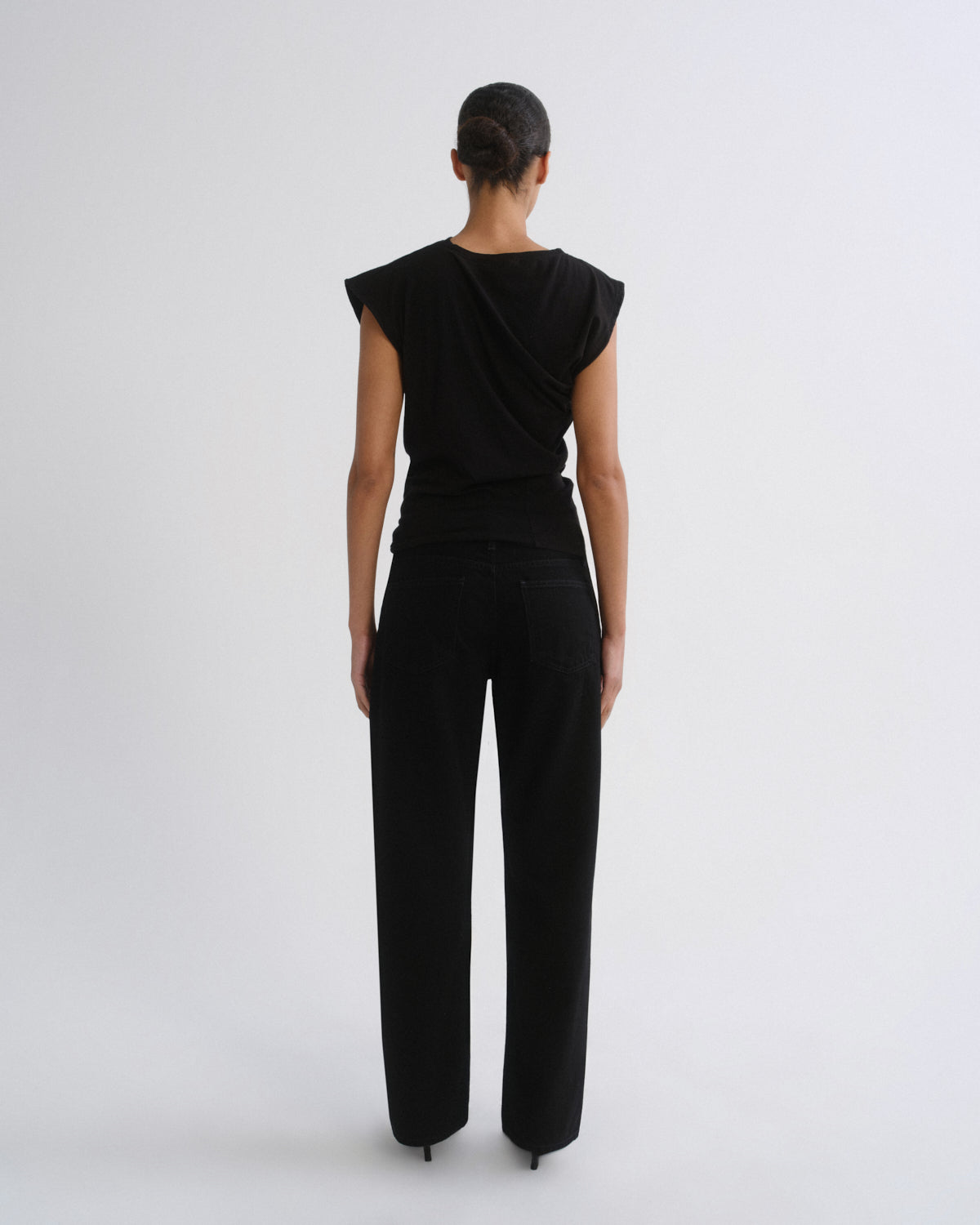 A model viewed from the back wearing a black draped sleeveless top and black wide-leg trousers against a light gray studio background.