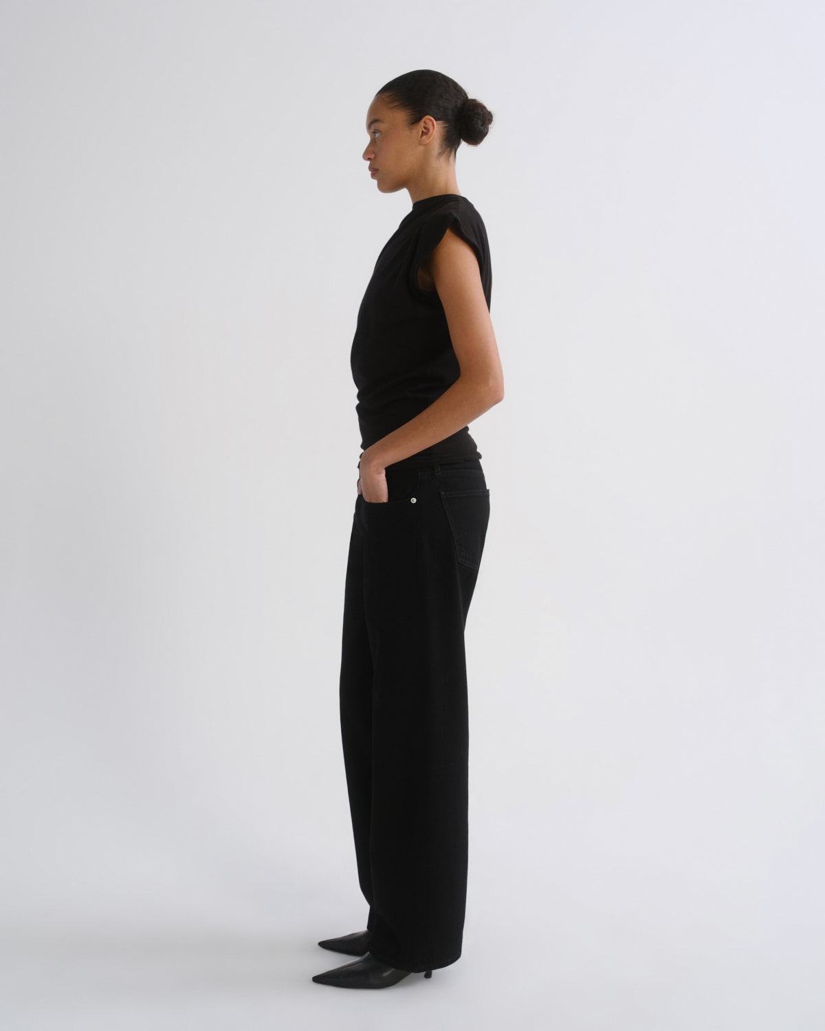 A model in side profile wearing a black sleeveless top and black wide-leg trousers, standing against a plain light gray studio background.