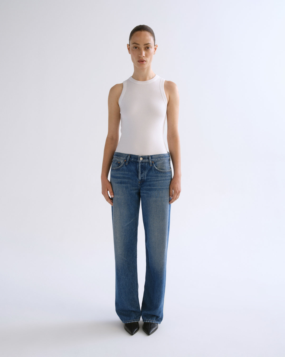 A model stands against a plain light gray studio background wearing a fitted sleeveless white top and blue wide-leg jeans.