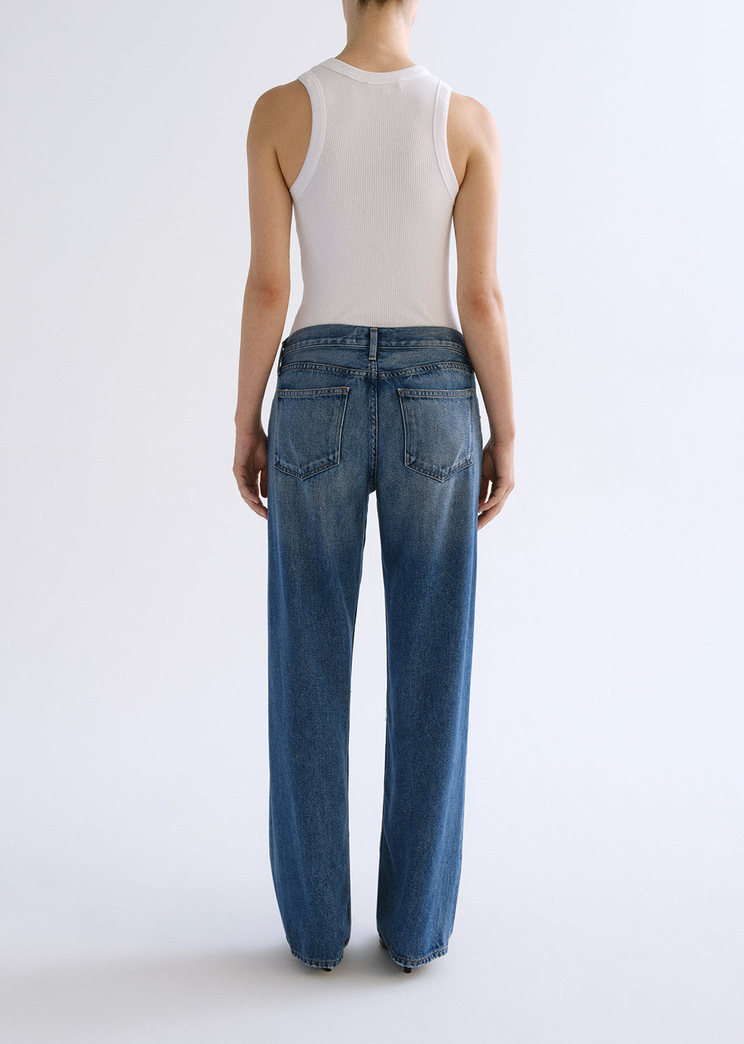 Person wearing white tank top with blue jeans on a grey background