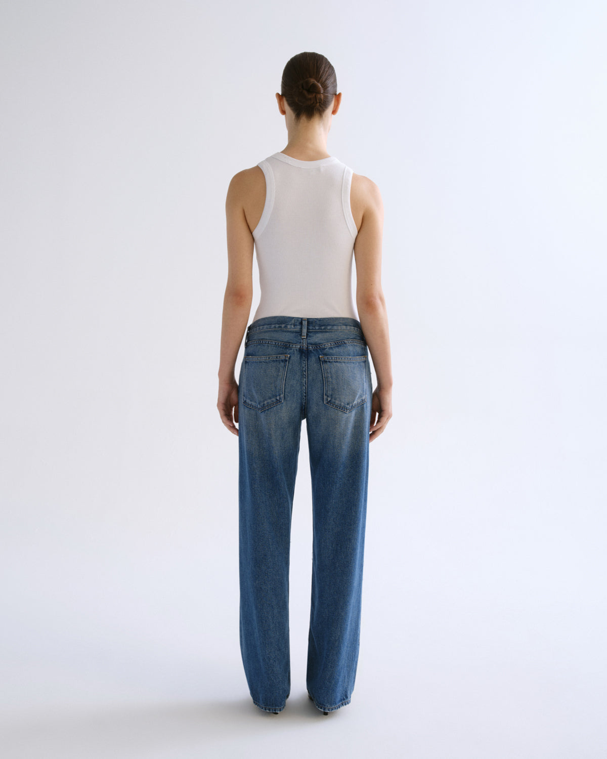A model shown from the back wearing a white ribbed tank top and wide-leg blue denim jeans against a plain white studio background.