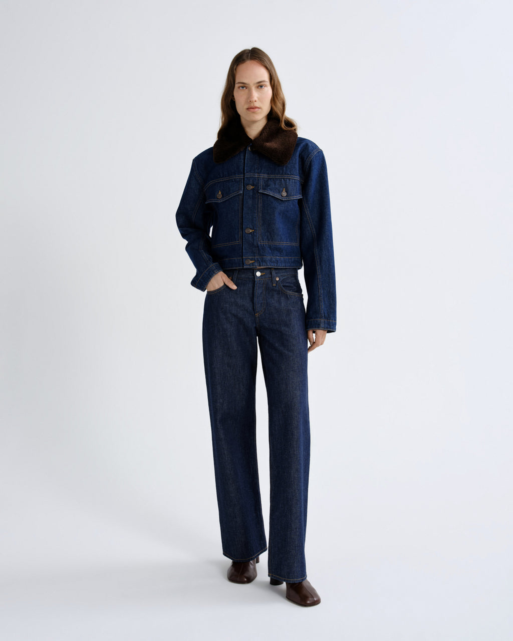 A model stands against a light gray studio background wearing a dark blue denim jacket with a brown collar and matching dark blue wide-leg denim pants.