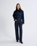 A model stands against a light gray studio background wearing a dark blue denim jacket with a brown collar and matching dark blue wide-leg denim pants.