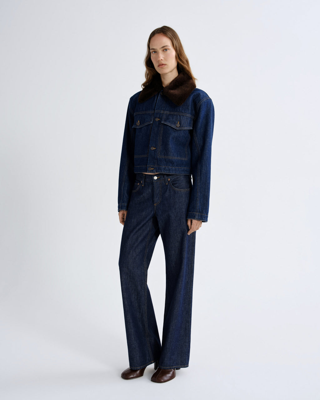 A model stands against a light gray studio background wearing a dark blue denim jacket with a dark brown fur collar, matching dark blue wide-leg denim pants, and brown leather shoes.
