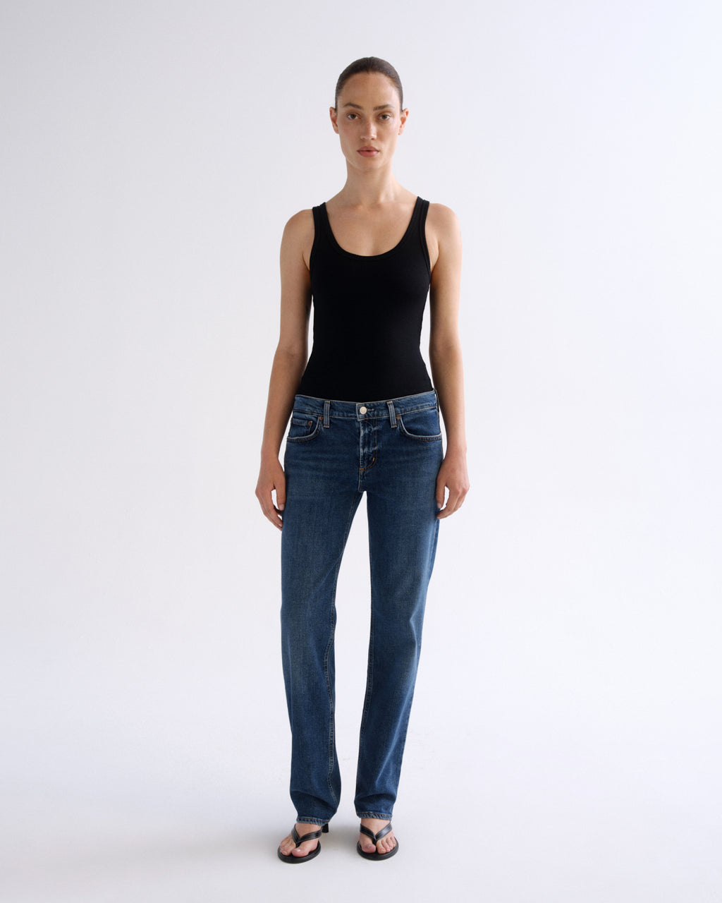 A model stands against a plain light gray studio background wearing a black sleeveless tank top, blue straight-leg jeans, and dark sandals.