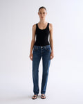 A model stands against a plain light gray studio background wearing a black sleeveless tank top, blue straight-leg jeans, and dark sandals.