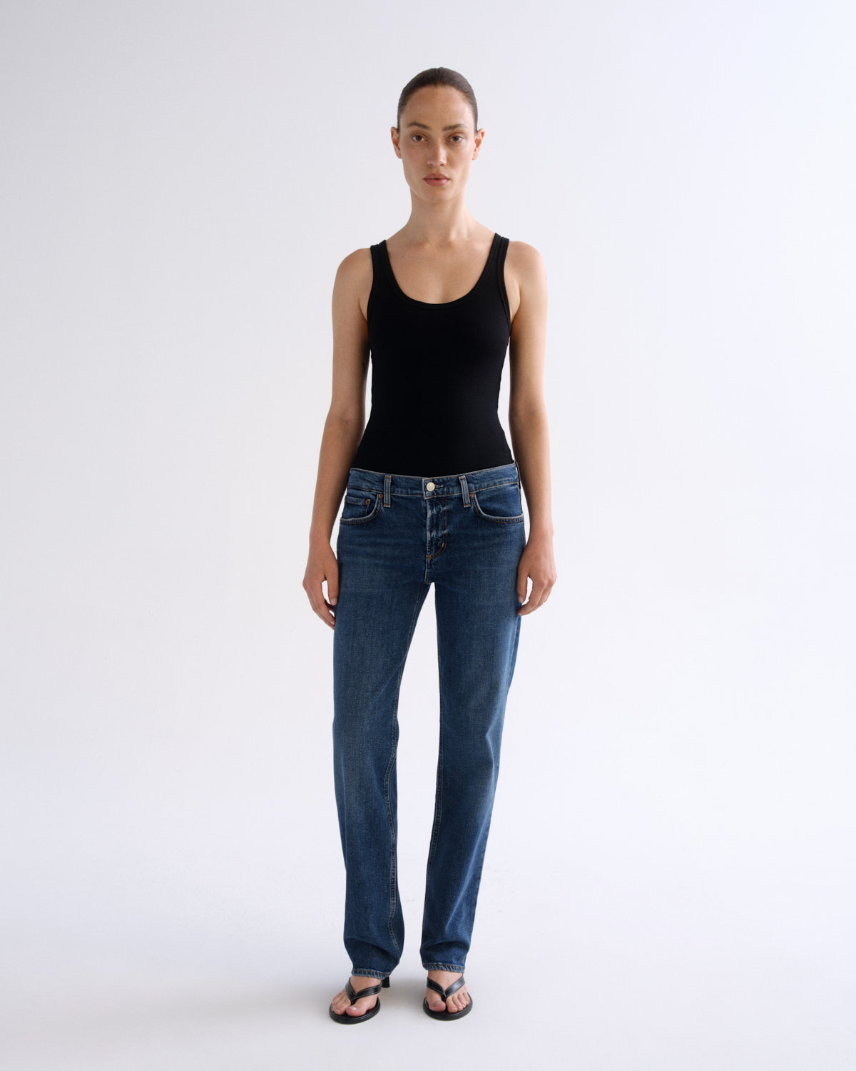 A model stands against a plain light gray studio background wearing a black sleeveless tank top, blue straight-leg jeans, and dark sandals.