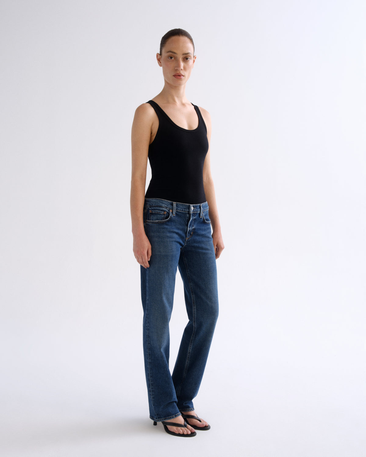 A woman modeling a black sleeveless tank top and blue straight-leg jeans against a plain light gray studio background.